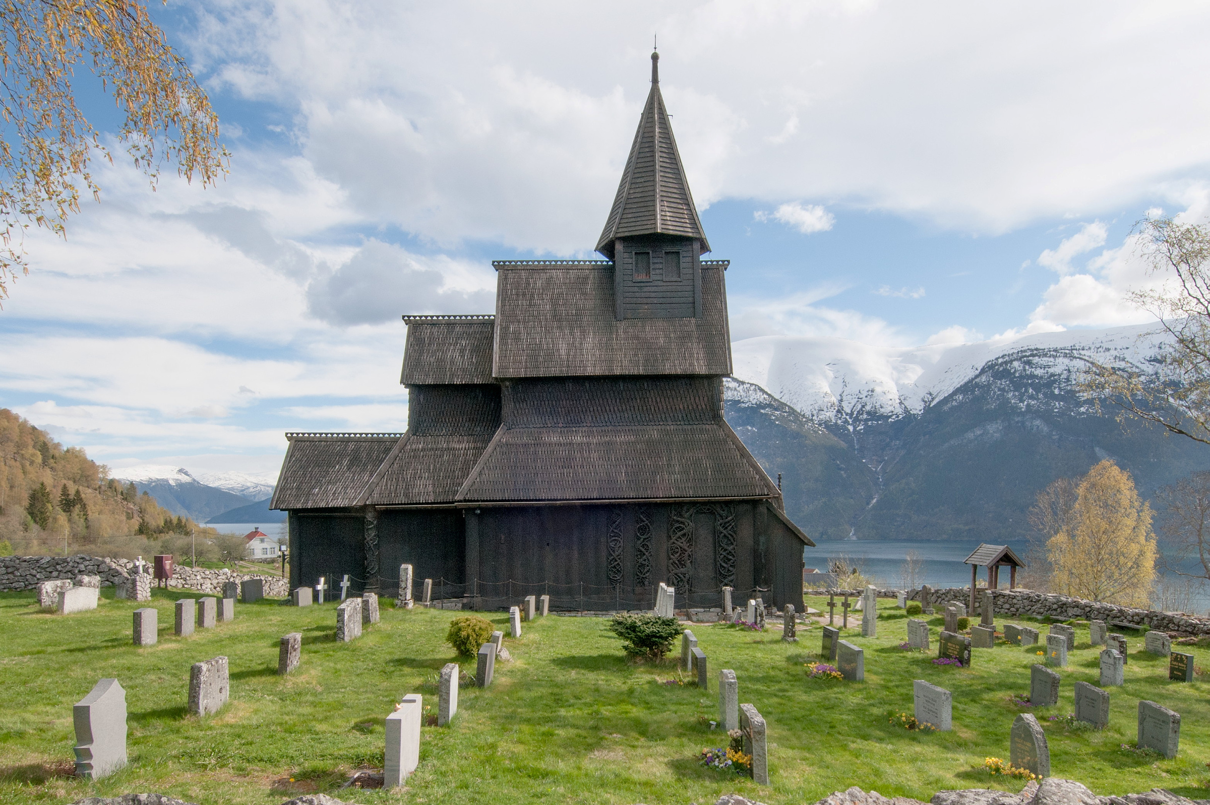 En stavkirke i tre med et høyt tårn i midten står på en kirkegård, omgitt av gravsteiner, med fjell og en innsjø i bakgrunnen.