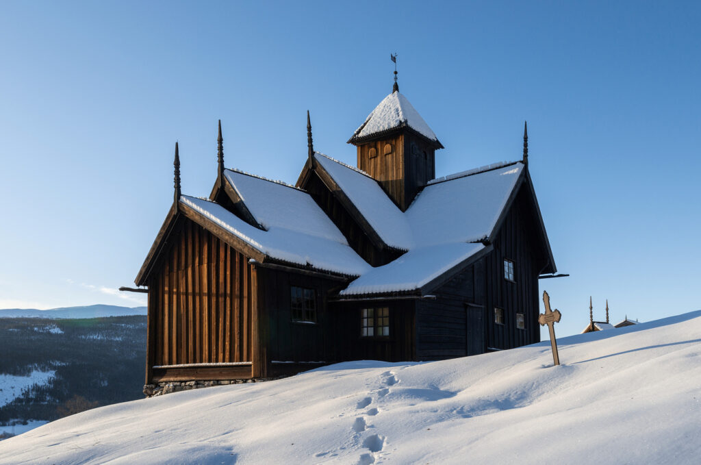 En stavkyrkjene i tre med bratte tak dekket av snø står i en snødekt skråning under en klarblå himmel, med fotspor som leder opp til inngangen.