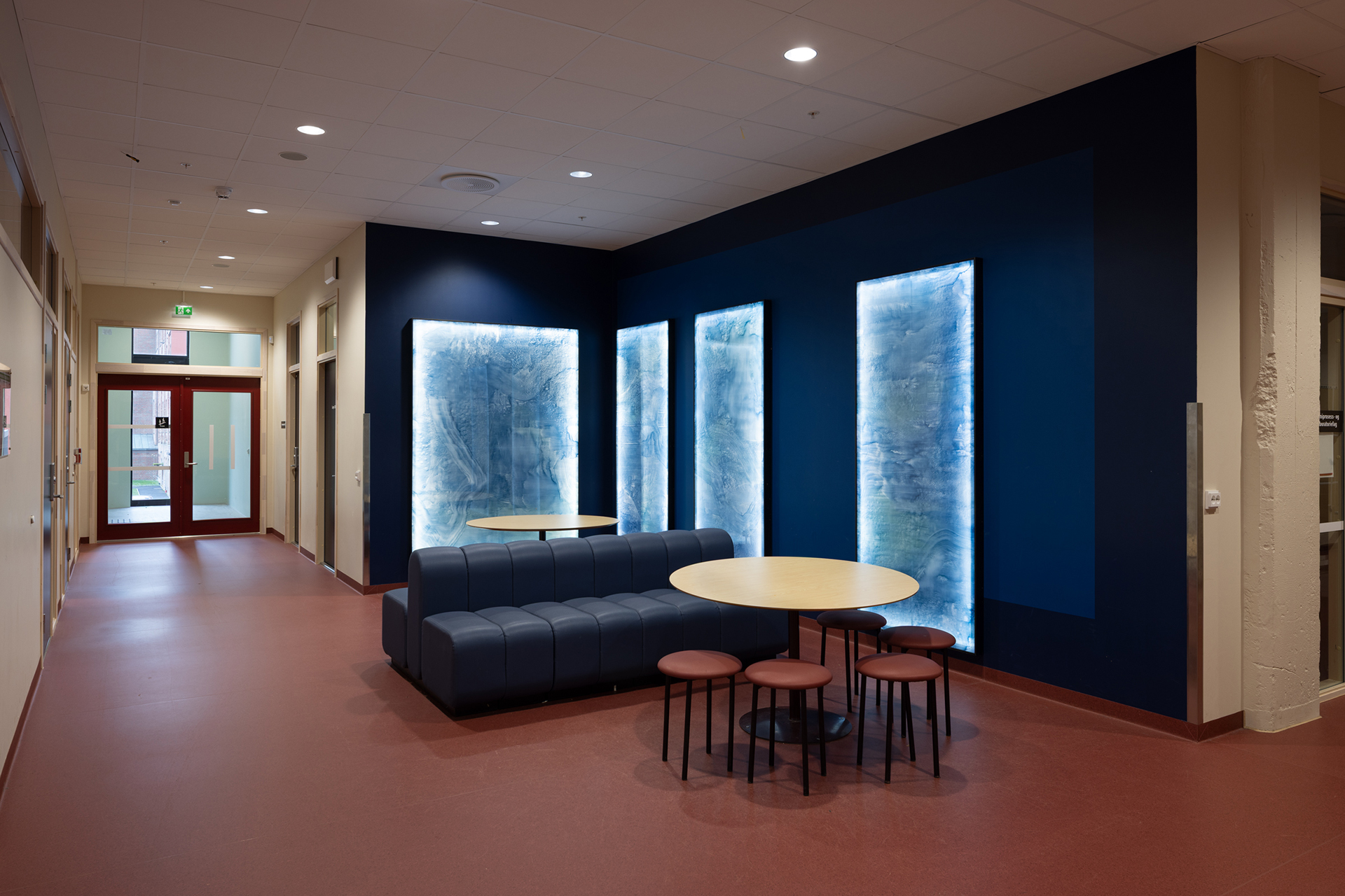 En moderne gang med sittegruppe, inkludert en blå sofa, runde bord og krakker, ved siden av opplyste veggpaneler med blått strukturmønster.