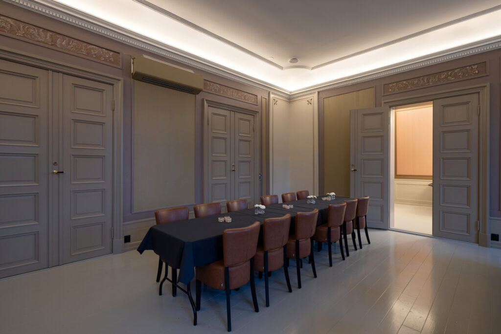 Stort rom med ornamenterte vegger, tak og eldre tredører med et langbord i midten med sort duk og brune skinnstoler.