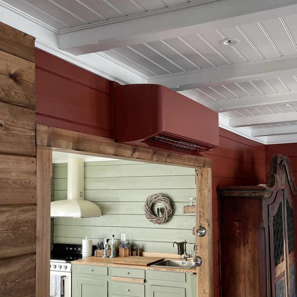 Et kjøkken med lysegrønne skap i tre, hvit kjøkkenvifte, tredetaljer og en rød veggmontert kanal over døren.