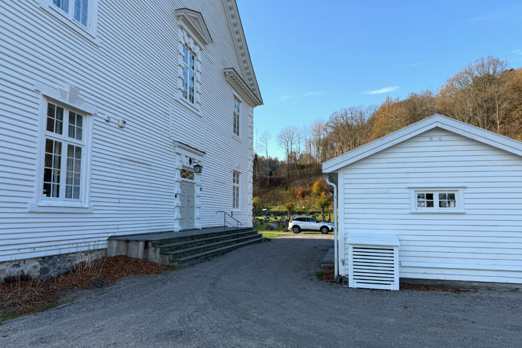 Stor hvit trebygning med trapper og rekkverk ved siden av et mindre hvitt skur, med en grussti, trær og en parkert bil i bakgrunnen under en klar himmel.
