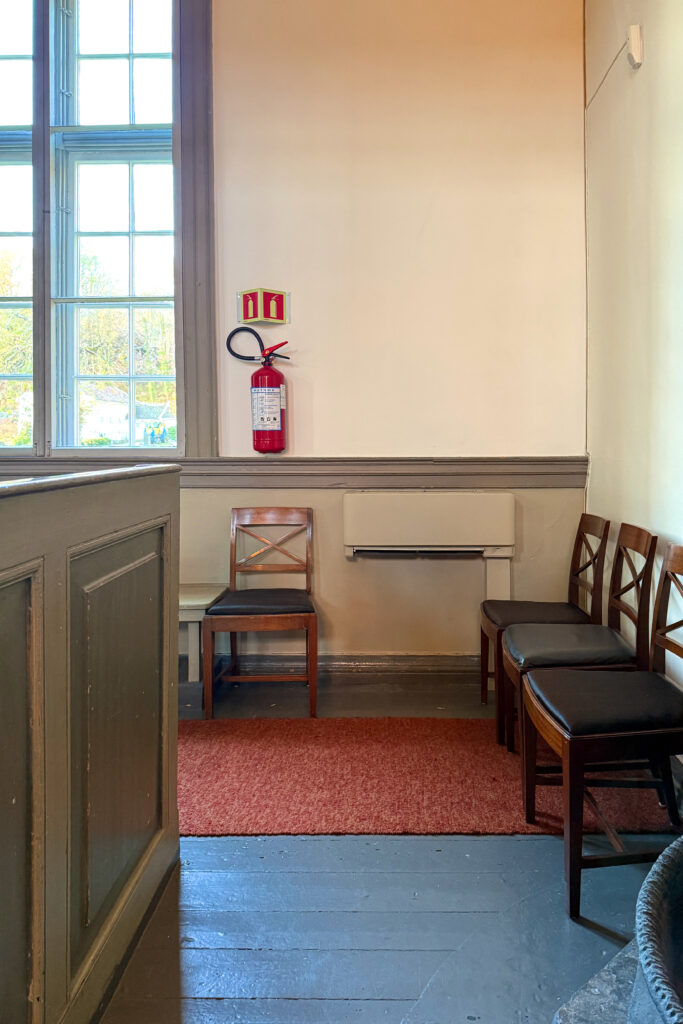 Et lite venteområde med trestoler, et rødt brannslukningsapparat på veggen og et stort vindu som slipper inn naturlig lys.