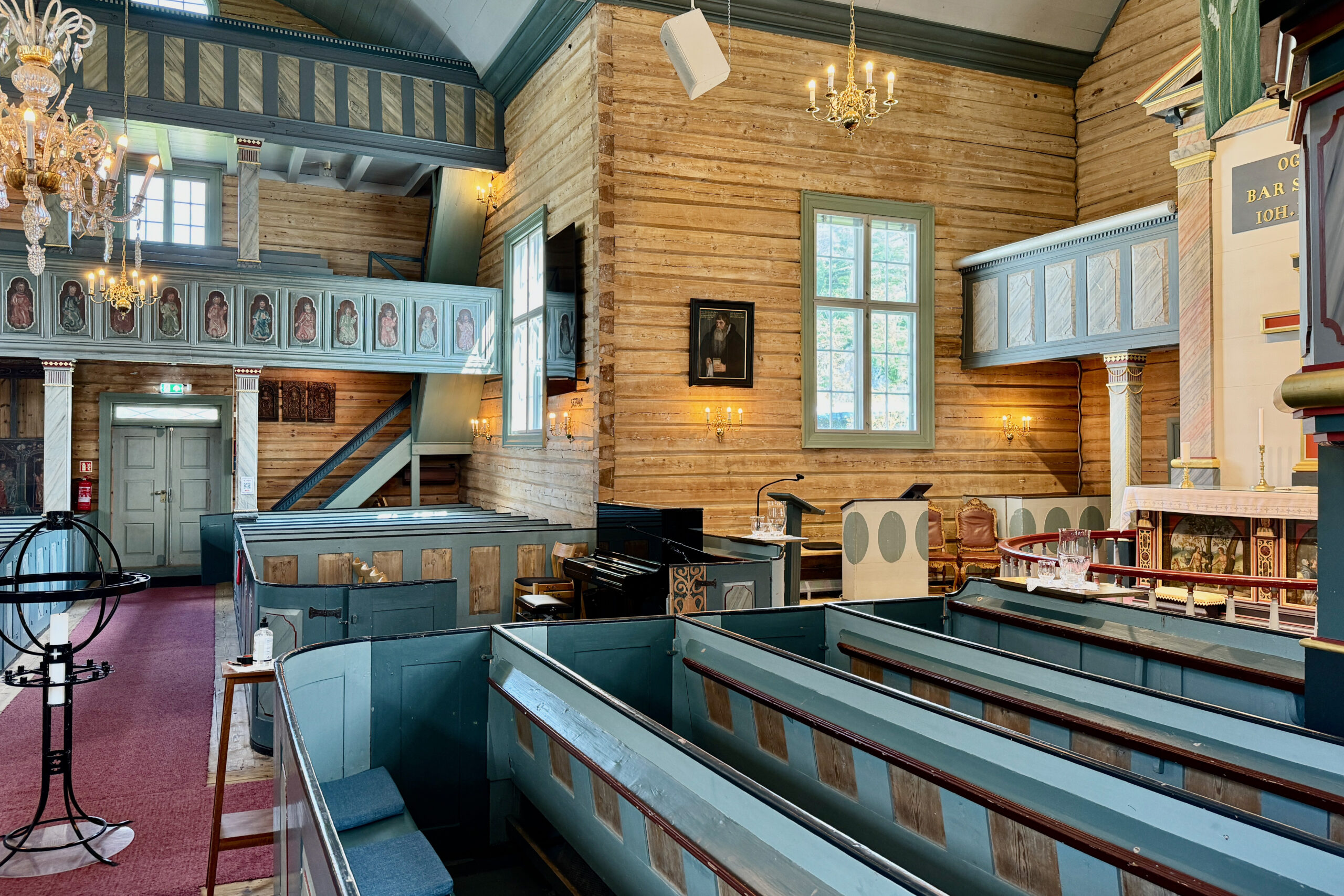 Interiør i en trekirke med blå kirkebenker, lysekroner, balkong og store vinduer som slipper inn naturlig lys.