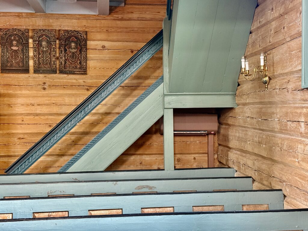 Interiør i en trebygning med blåmalte kirkebenker, en trapp, veggmonterte lysestaker og tre dekorative plaketter på en tømmervegg.