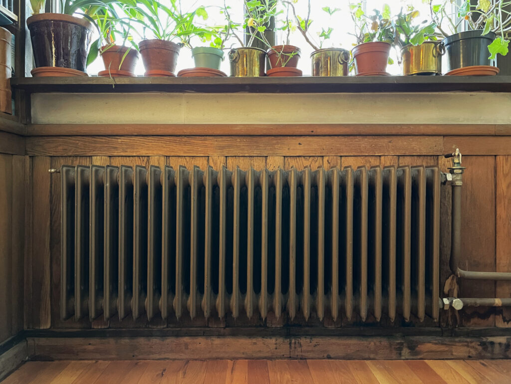 En gammel metallradiator under en trehylle med flere potteplanter, med sollys som faller inn gjennom et vindu bak dem.