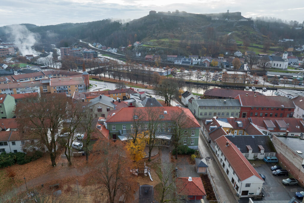 Flyfoto av en liten by med røde takbygninger, en elv og en festning på en bakketopp i bakgrunnen på en overskyet dag.