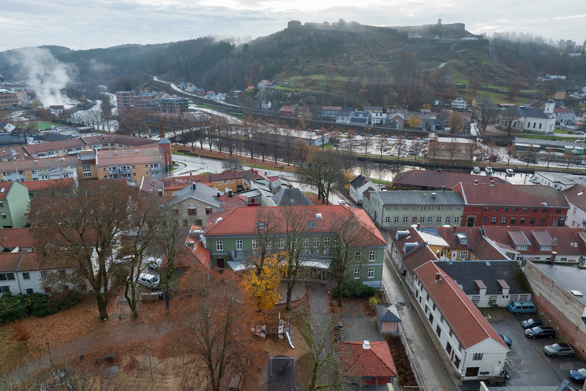 Flyfoto av en liten by med røde takbygninger, en elv og en festning på en bakketopp i bakgrunnen på en overskyet dag.