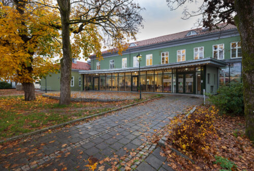 Eksempelsamling 2 En asfaltert gangvei fører til en moderne glassinngang i en grønn museumsbygning, omgitt av høsttrær og løvfall.