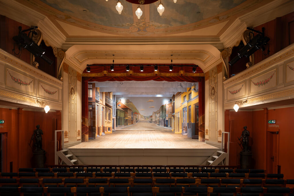 Utsmykket teaterinteriør med tomme seter som vender mot en scene med en malt gatekulisse, innrammet av dekorative vegger og tak.