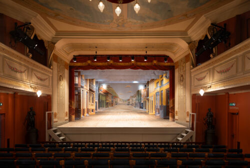 Eksempelsamling 1 Utsmykket teaterinteriør med tomme seter som vender mot en scene med en malt gatekulisse, innrammet av dekorative vegger og tak.