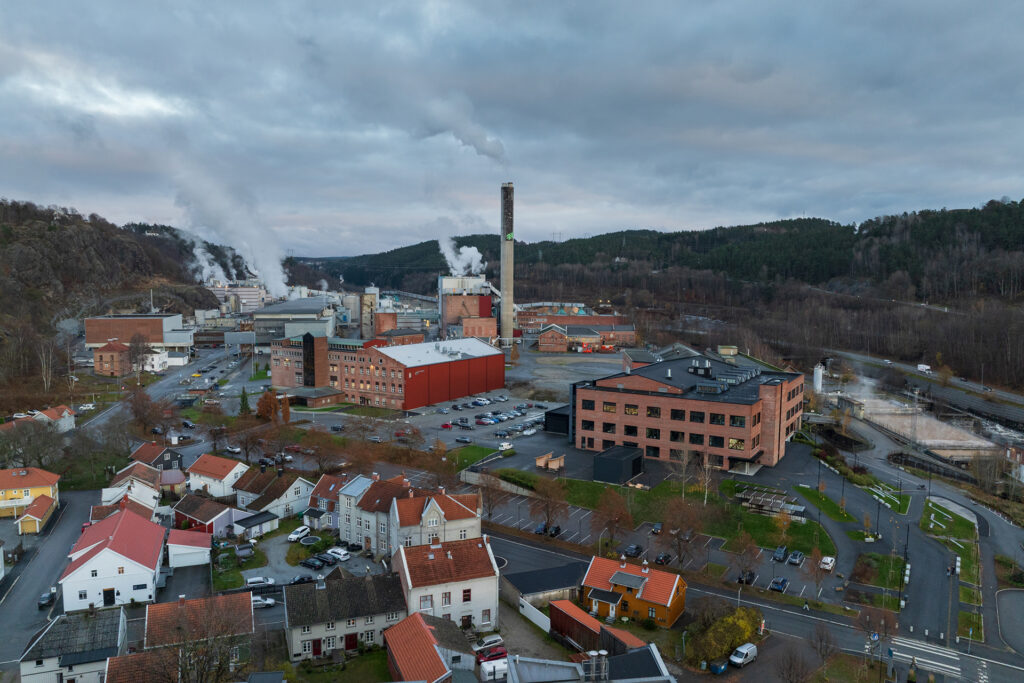 Flyfoto av et industrikompleks med rykende skorsteiner, omgitt av bolighus, veier og skogkledde åser under en overskyet himmel.