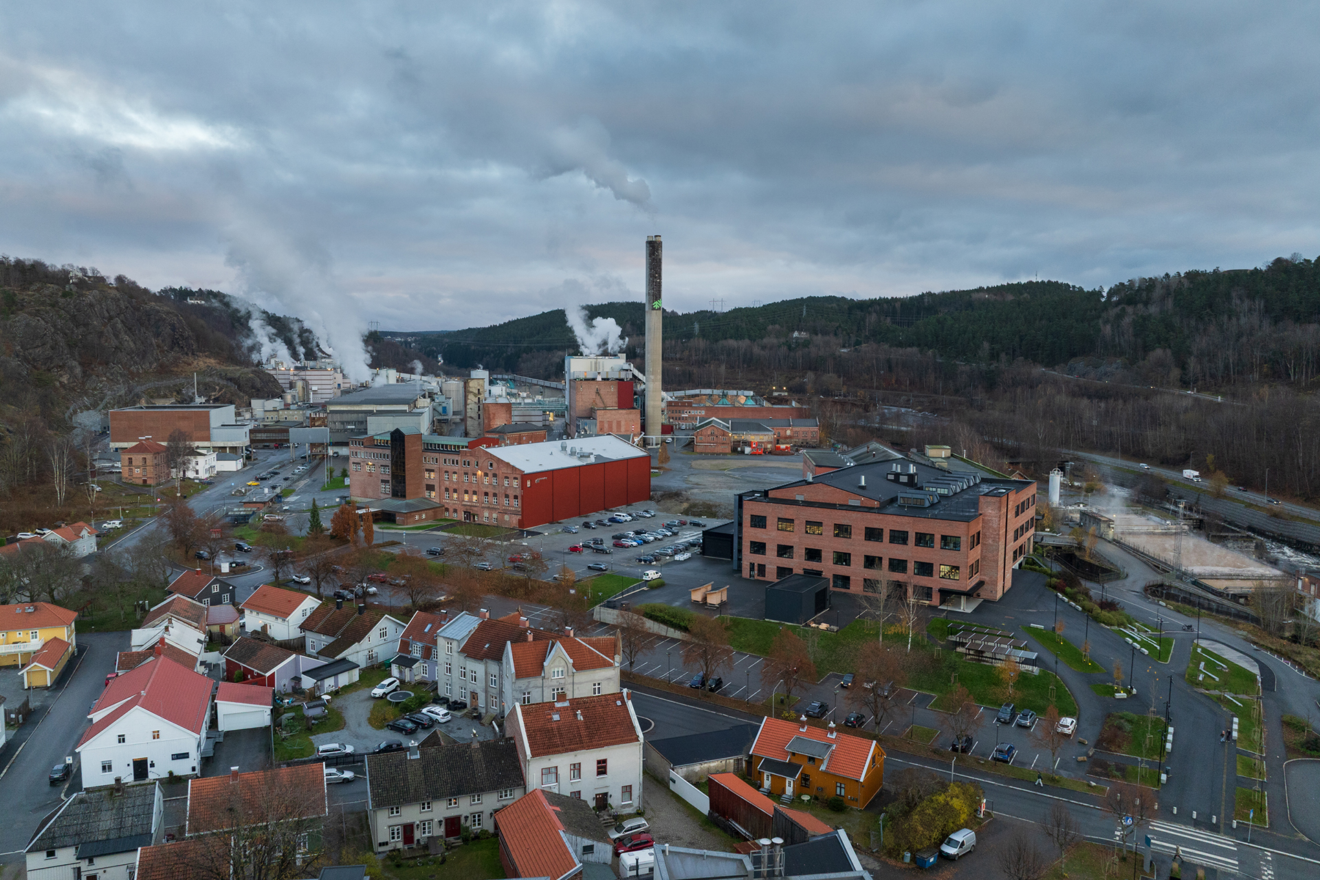 Flyfoto av et industrikompleks med rykende skorsteiner, omgitt av bolighus, veier og skogkledde åser under en overskyet himmel.