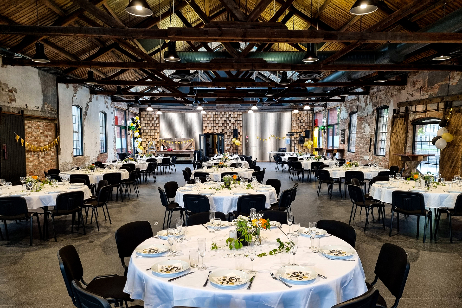 På rett spor i Byparken 8 En dekorert hall i industristil med runde bord dekket til et arrangement, med hvite duker, blomsteroppsatser og svarte stoler. Ballonger og girlandere er hengt opp rundt i rommet.