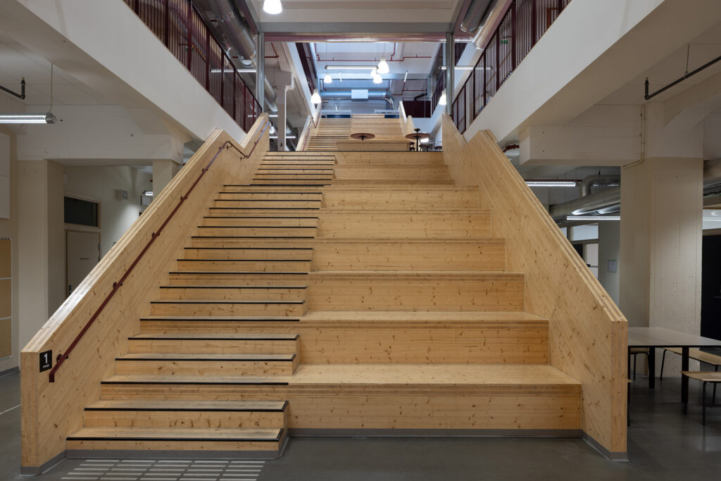 Bred tretrapp med integrerte sitteplasser i et moderne, industrielt bygningsinteriør, opplyst av taklamper og omgitt av åpne korridorer og rekkverk.