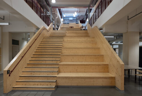 Eksempelsamling 4 Bred tretrapp med integrerte sitteplasser i et moderne, industrielt bygningsinteriør, opplyst av taklamper og omgitt av åpne korridorer og rekkverk.