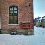 En murbygning med et grønt innrammet vindu og en ventilasjonsåpning ligger ved siden av en snødekt gate; i bakgrunnen skimtes bolighus.