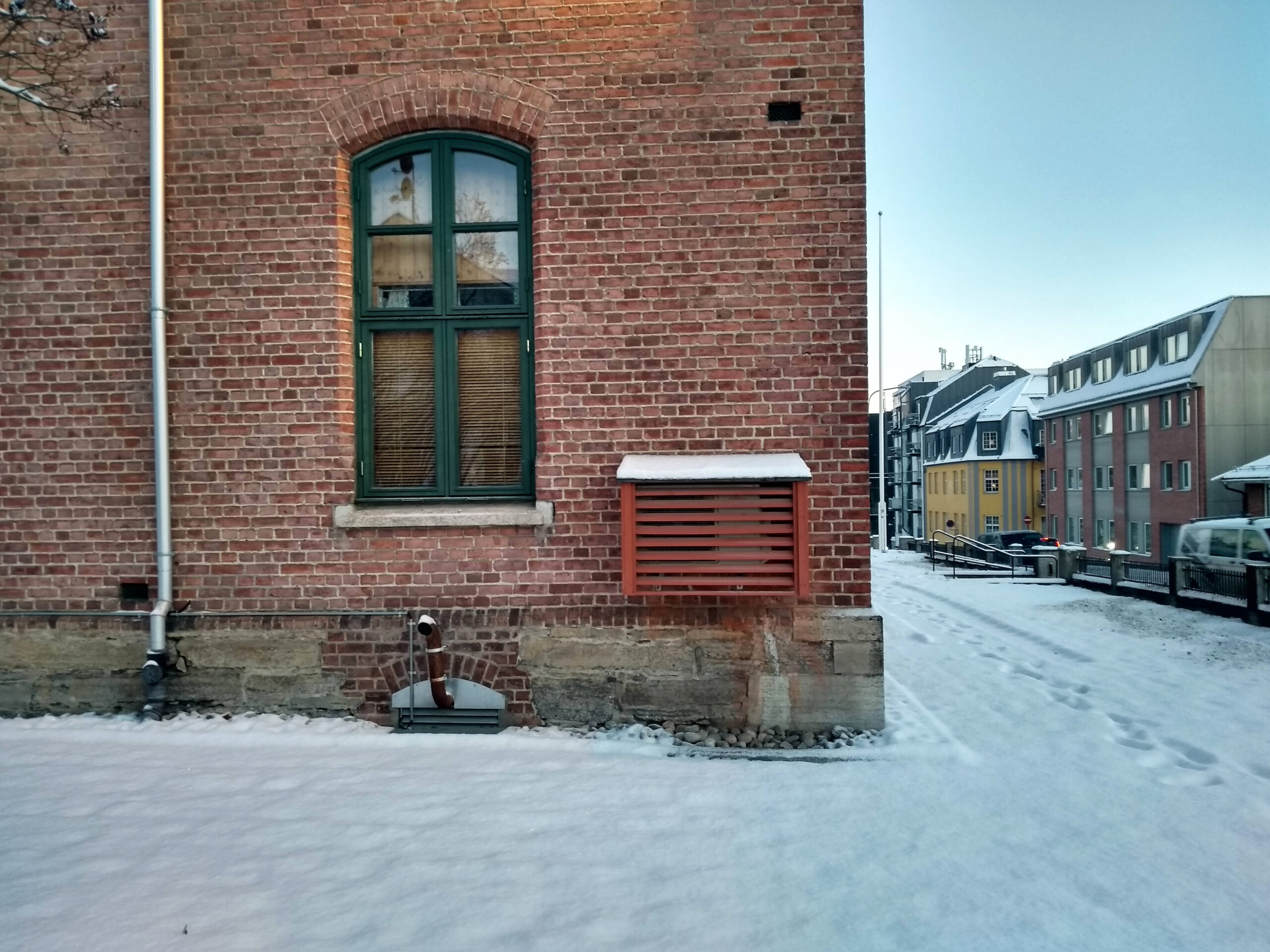 En murbygning med et grønt innrammet vindu og en ventilasjonsåpning ligger ved siden av en snødekt gate; i bakgrunnen skimtes bolighus.