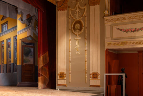 Eksempelsamling 2 En teaterscene med et malt bakteppe av et gult hus, utsmykkede veggdekorasjoner og en balkong med blomstermotiver.