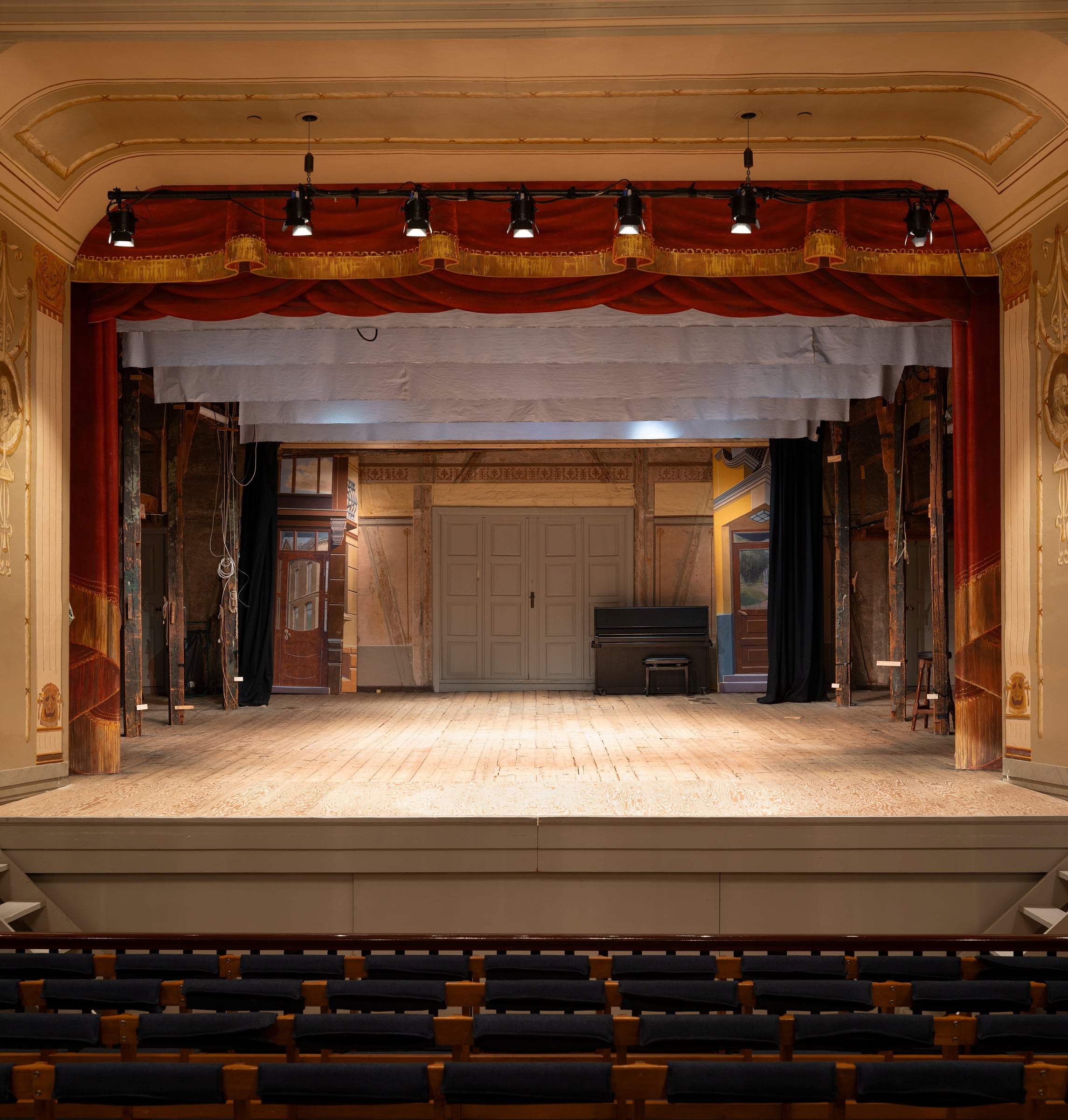Tryggleik i kulissane 6 Tom teaterscene med tregulv, røde gardiner, scenelys og rader med tomme seter i forgrunnen.