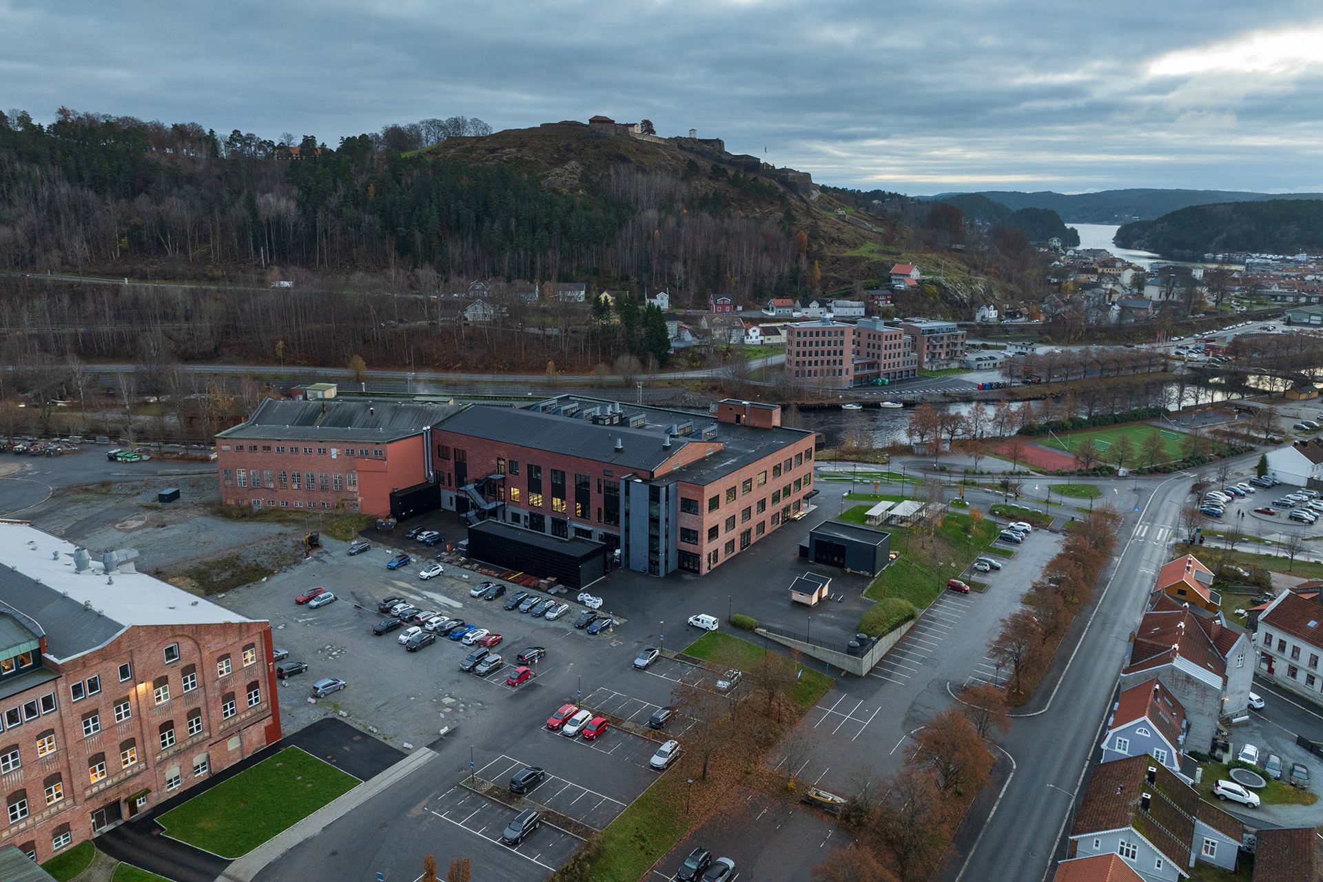 Flyfoto av en campus med flere mursteinsbygninger, en parkeringsplass med biler, en ås med trær i bakgrunnen og en elv synlig til høyre.