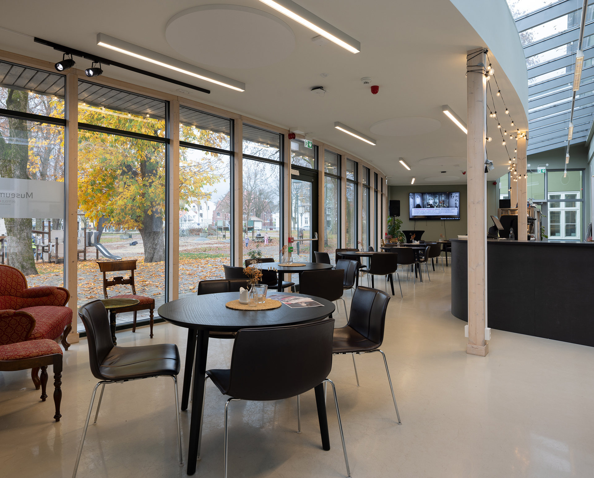 Paleet som opna seg for parken, og for byen 8 Moderne kaféinteriør med svarte bord og stoler, store vinduer som viser høsttrærne utenfor, og en disk i bakgrunnen.