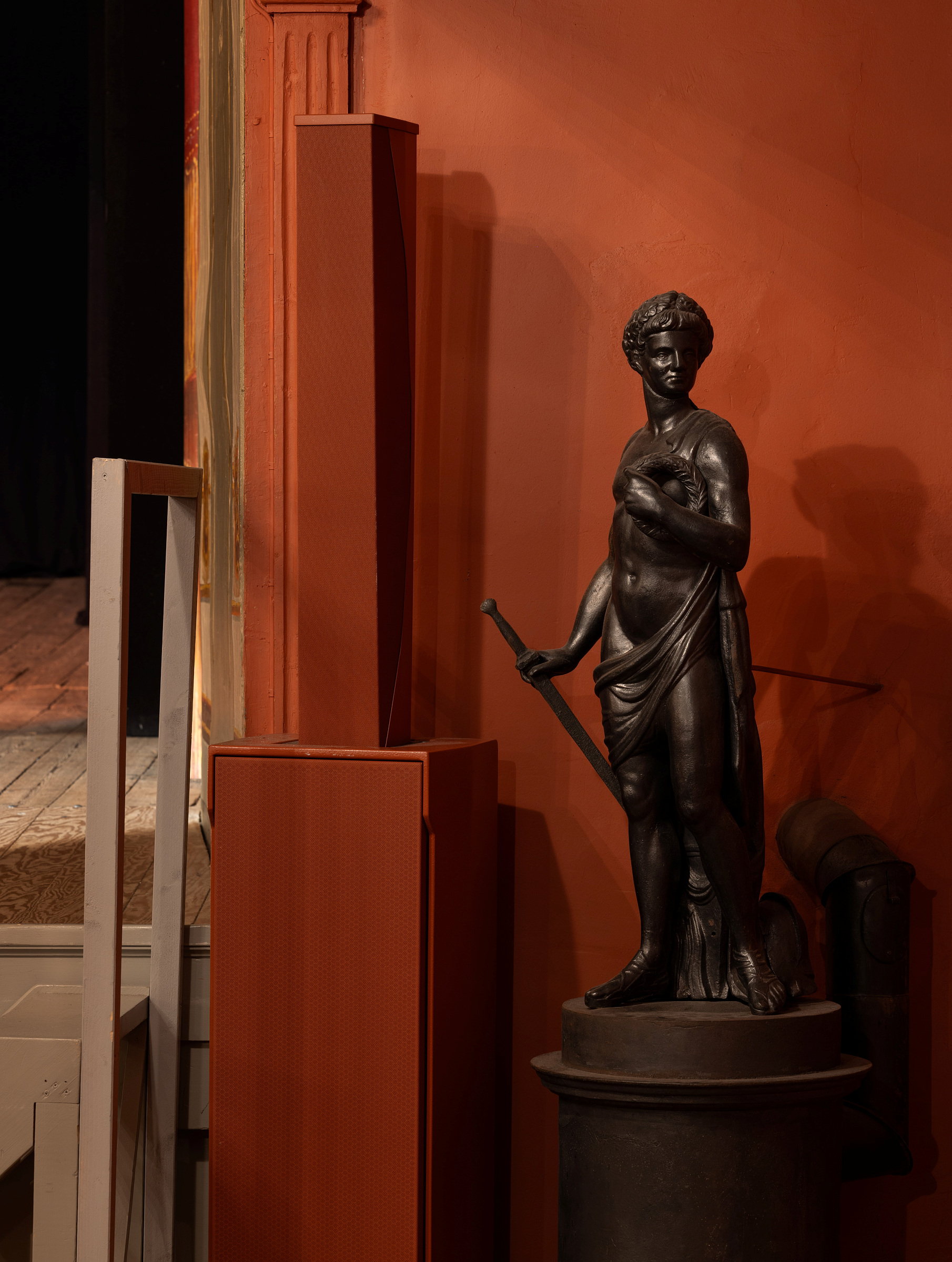 Rigga med både tradisjonelle og moderne løysingar 8 En bronsestatue av en delvis drapert kvinne som holder en stav, står på en sokkel mot en rød vegg nær en scene av tre.