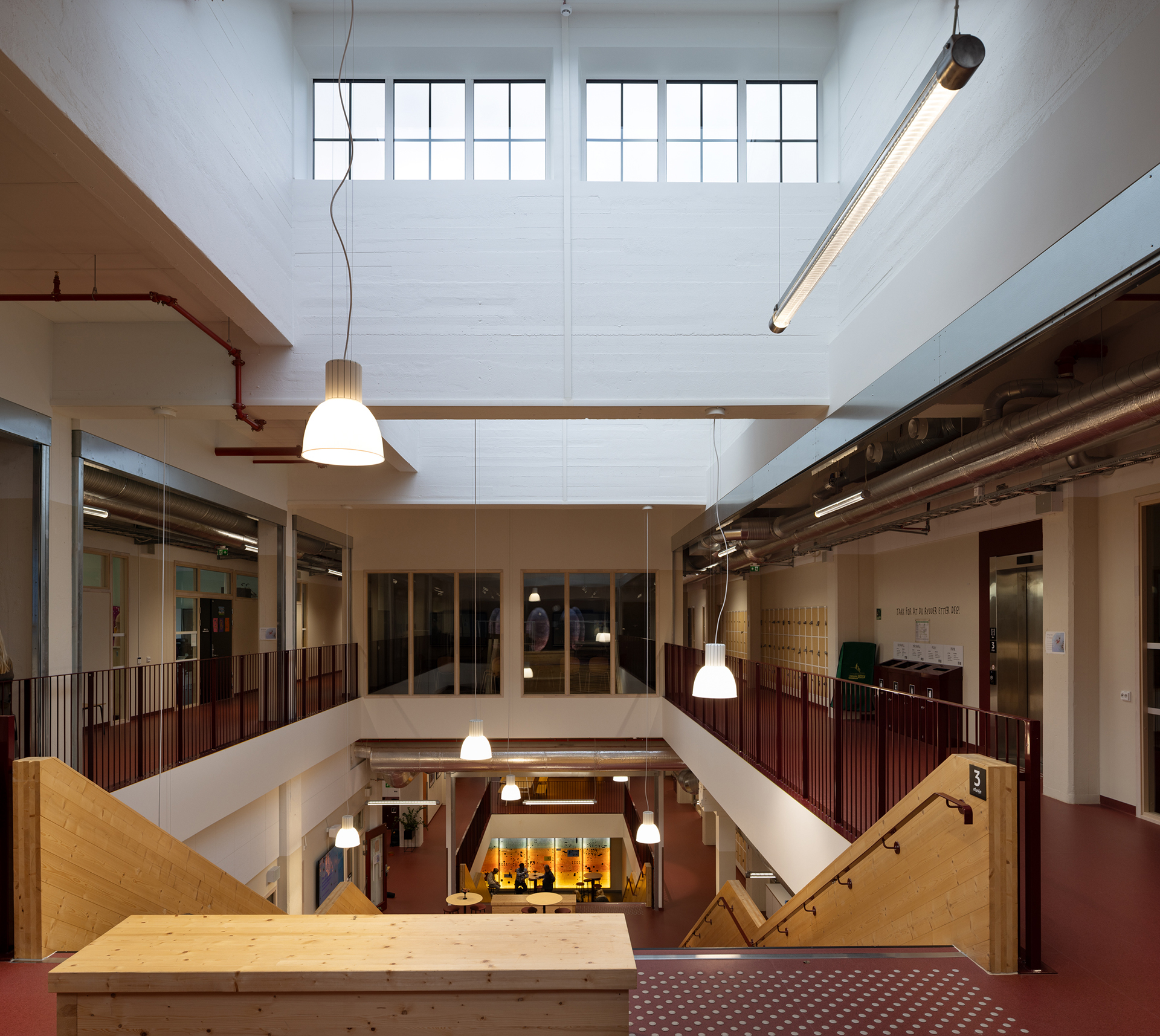 Et moderne, åpent interiør med stor takhøyde, store vinduer, trapper og hengende lamper, sett fra et øvre plan med utsikt over området nedenfor.