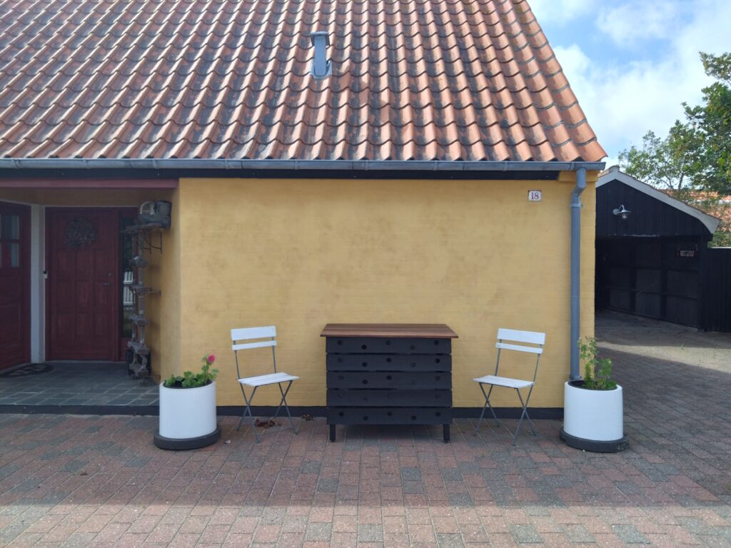 En gul husvegg med tegltak, to hvite stoler, et svart bord og to store hvite krukker på en mursteinsbelagt uteplass.