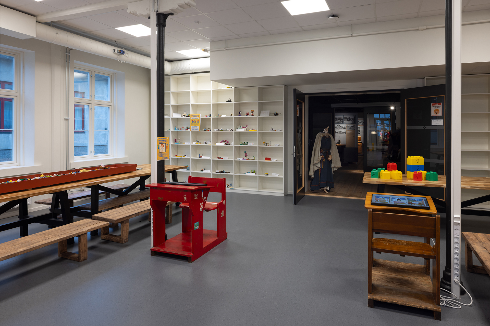 Kameleonen ved parken 9 Rom med trebenker, en LEGO-byggestasjon, tomme, hvite hyller og en døråpning som fører til et utstillingsområde; vinduer langs venstre vegg.