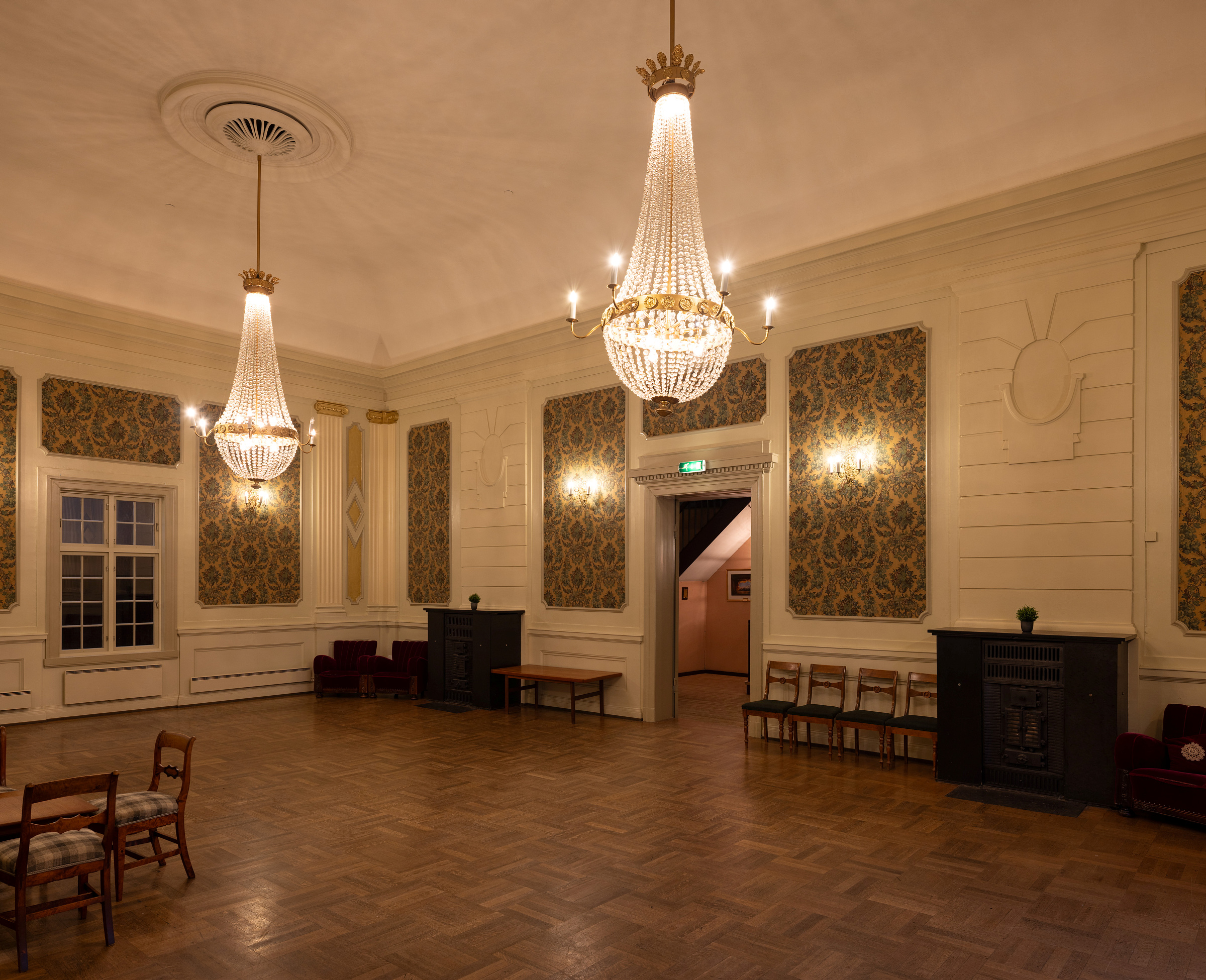 Stor, tom ballsal med utsmykkede lysekroner, mønstret tapet, tregulv og noen få stoler langs veggene.