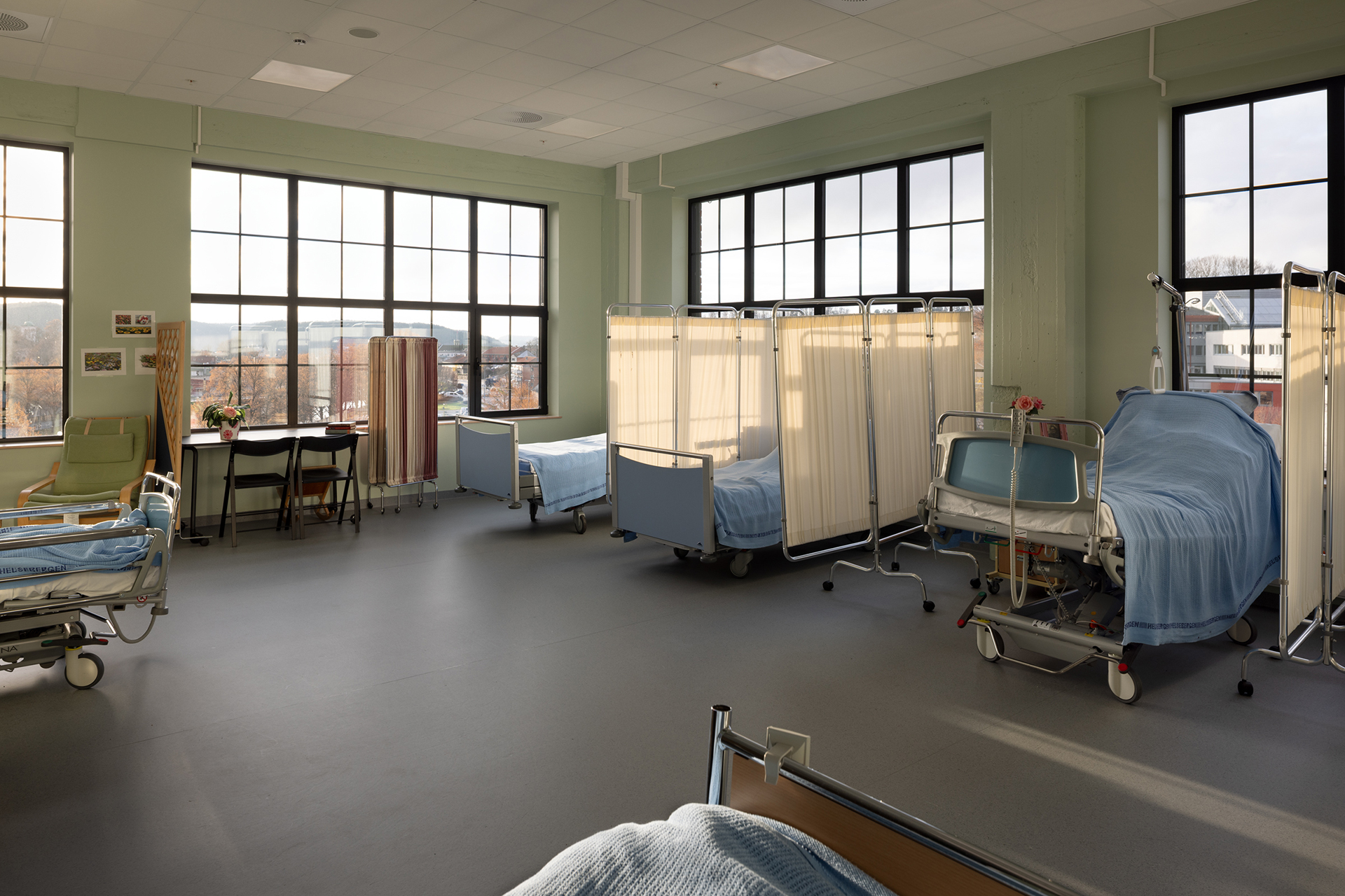 En sykehusavdeling med flere tomme senger, innsynsgardiner, store vinduer og naturlig lys som fyller rommet.