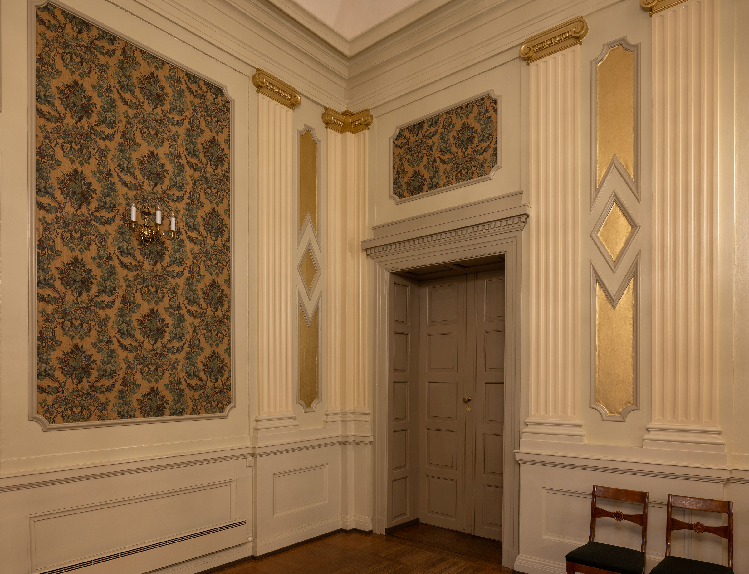 Utsmykket romhjørne med beige panelvegger, gullaksenter, blomstrete tapetpaneler, en tredør og to trestoler mot veggen.
