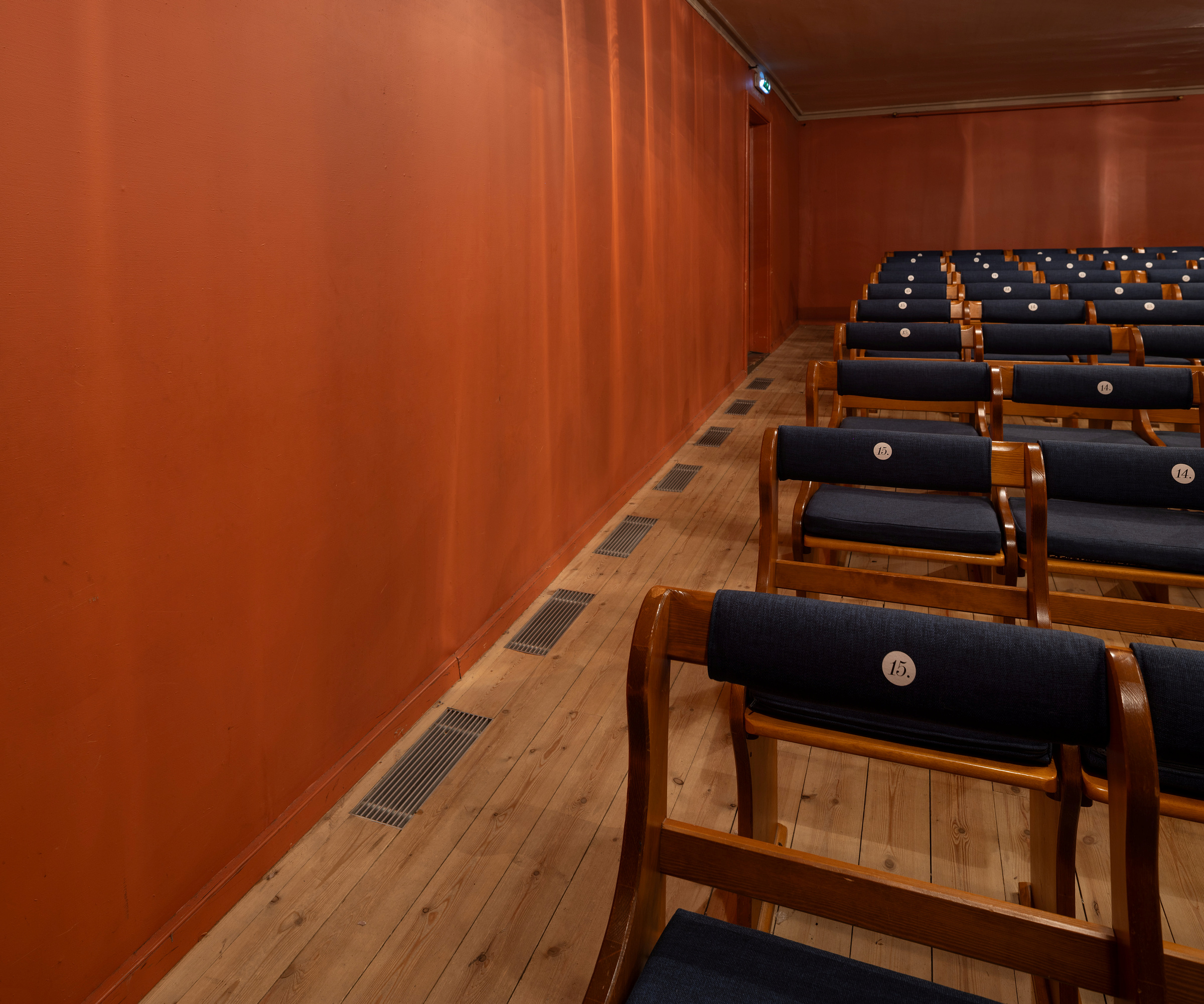 Rigga med både tradisjonelle og moderne løysingar 9 I et rom med oransje vegger og tregulv står rekker av trestoler med marineblå puter.