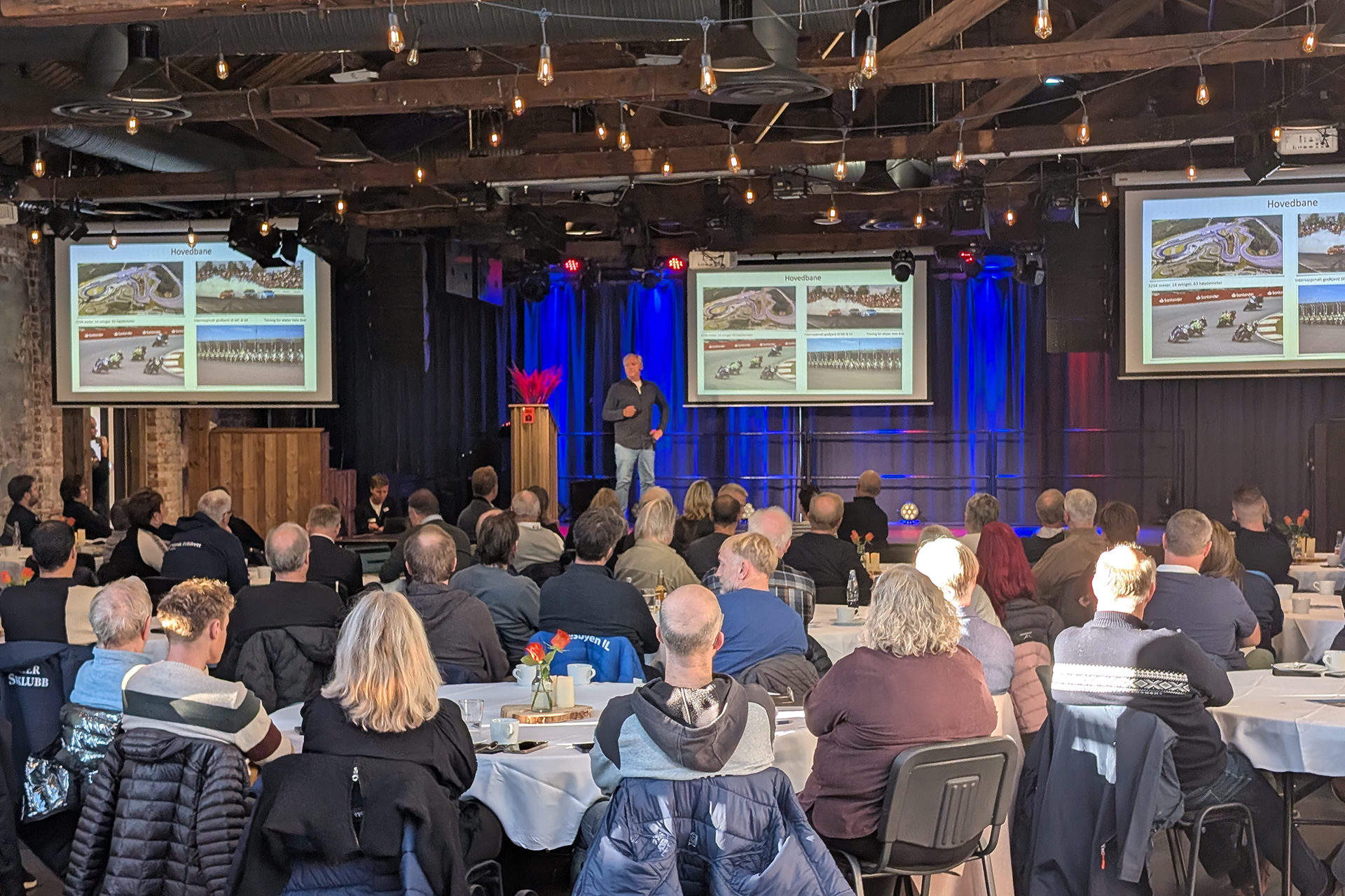 På rett spor i Byparken 9 En mann holder en presentasjon med lysbilder som viser travbaner for et publikum som sitter ved bordene i et rom med synlige bjelker og lyskastere.