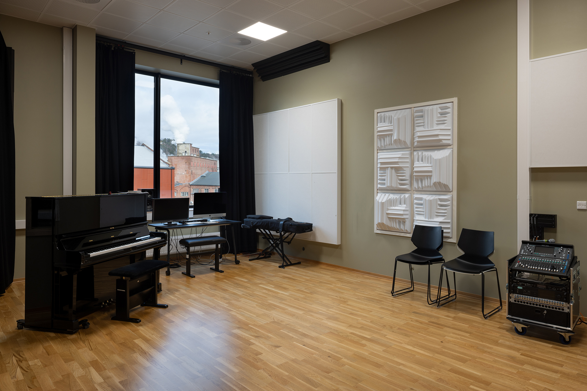 Et musikkklasserom med piano, musikkutstyr, stoler, akustiske paneler på veggene og et stort vindu med utsikt over bygningene utenfor.