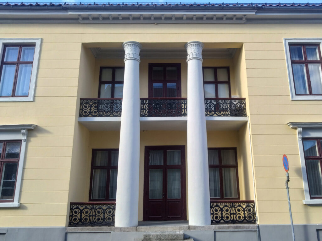 Gul fasade med to store hvite søyler, to balkonger med utsmykkede rekkverk og symmetriske vinduer. Et skilt med parkeringsforbud er synlig til høyre.