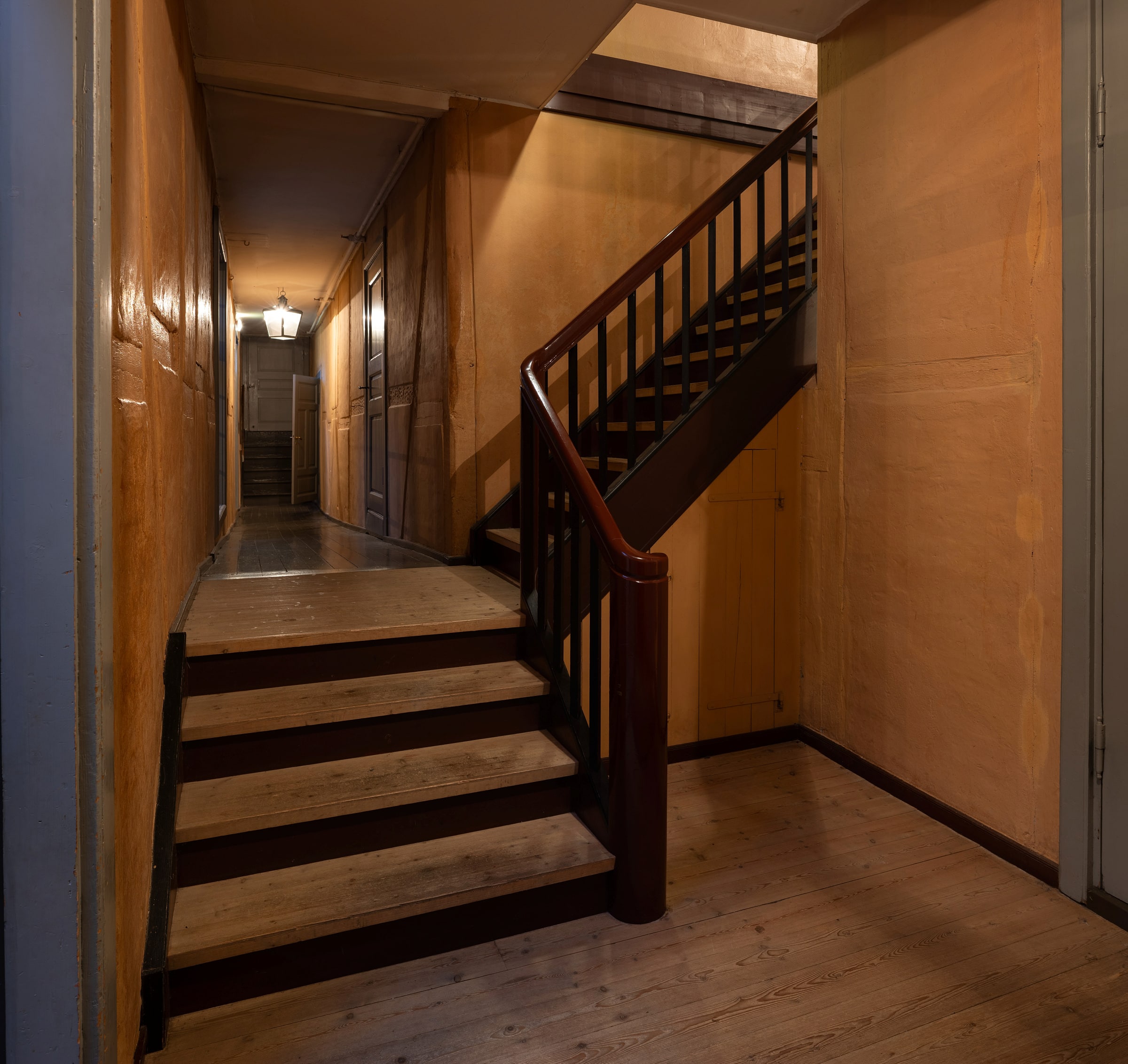 Tryggleik i kulissane 10 Svakt opplyst gang med tregulv og en trapp som fører oppover; dører og vegger er malt i brune nyanser.