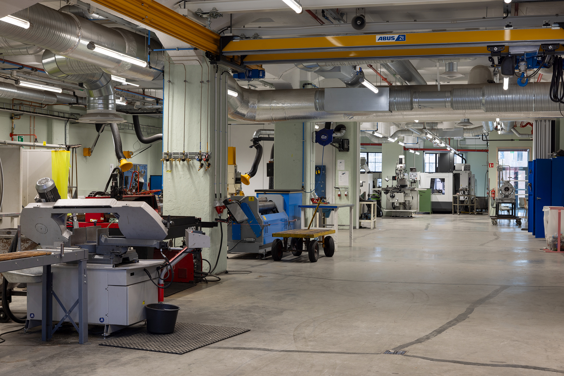 Romslig industriverksted med ulike metallbearbeidingsmaskiner, arbeidsbenker, traverskran og ventilasjonskanaler, godt opplyst av naturlig og kunstig lys.