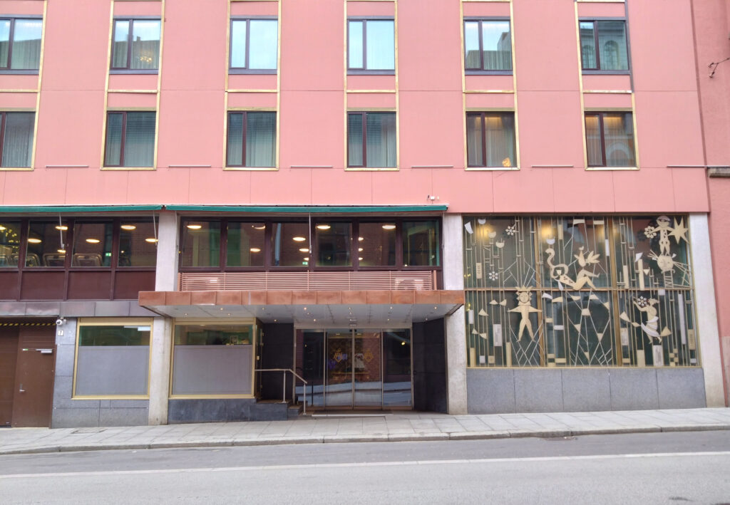 En rosa bygning med store vinduer, inngangsparti i glass og dekorative metallarbeider på høyre side, sett fra den andre siden av en tom gate.