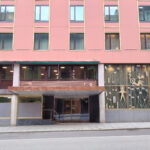 En rosa bygning med store vinduer, inngangsparti i glass og dekorative metallarbeider på høyre side, sett fra den andre siden av en tom gate.