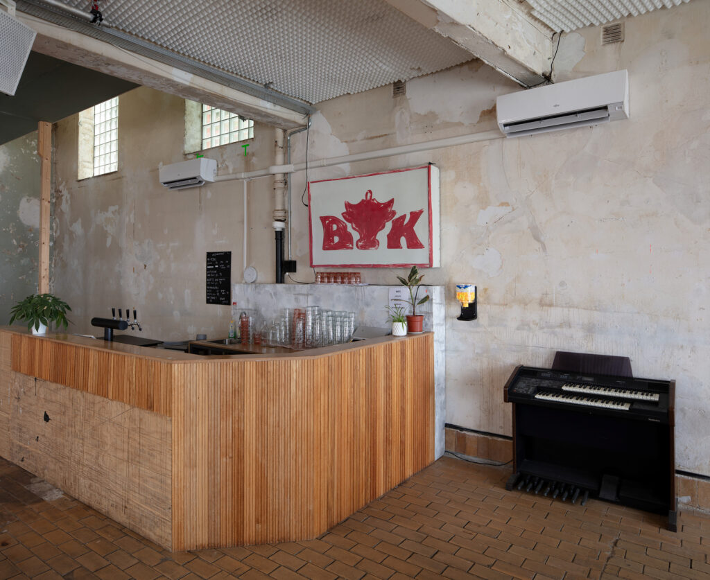 En bar med en disk i tre, flere glass, en liten meny, en plante, et elektrisk orgel og et veggskilt med et rødt oksehode og bokstavene BOK.