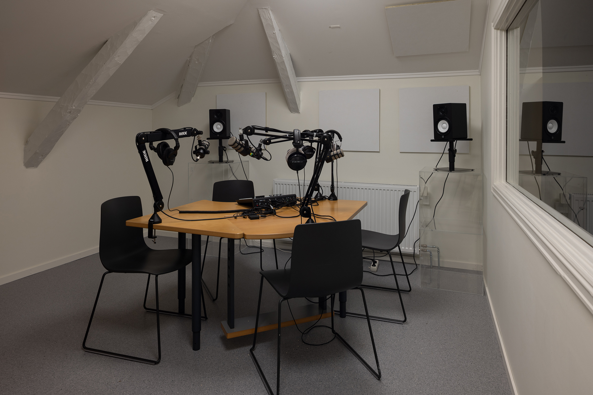 Paleet som opna seg for parken, og for byen 10 Et podkaststudio med fire stoler rundt et bord, mikrofoner på justerbare armer, hodetelefoner og høyttalere på stativer i et lydisolert rom.