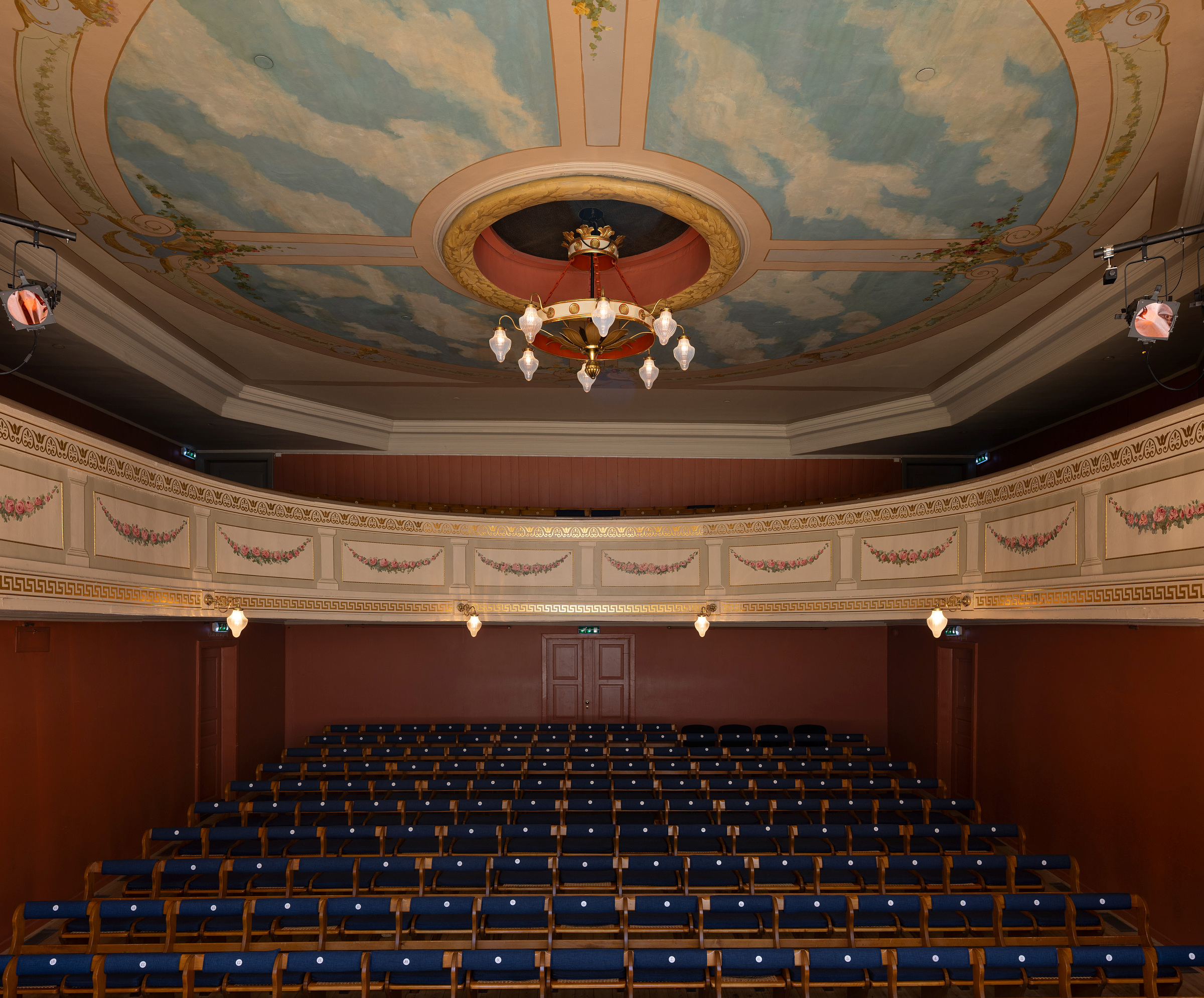 Et tomt teater med rader av blå seter, en utsmykket balkong og et malt tak med skyer og en lysekrone.