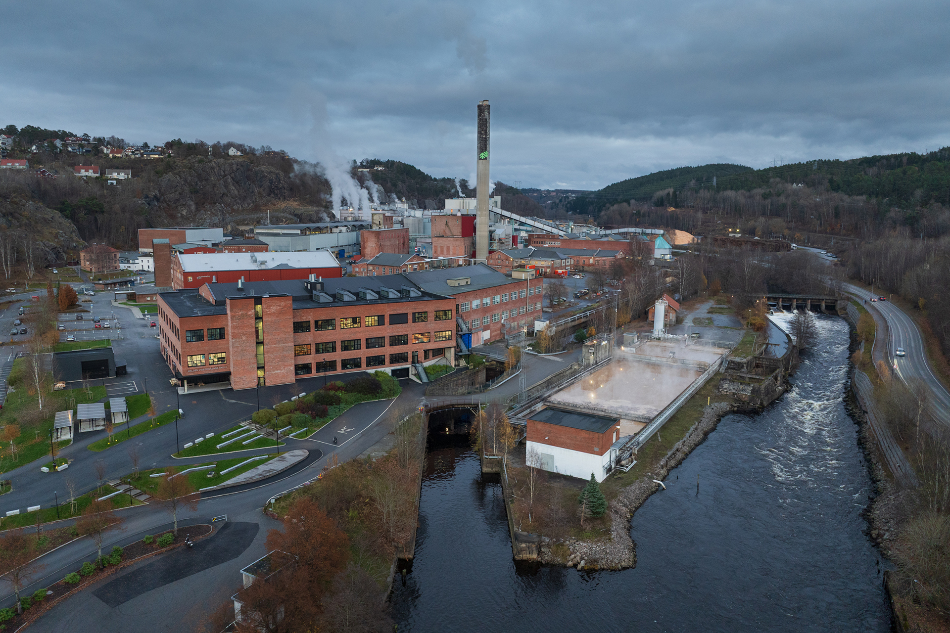 Skulen knyt industrien og byen i hop 6 Et stort industrianlegg med mursteinsbygninger, en høy skorstein og damputslipp ligger ved en elv, med veier og skogkledde åser i bakgrunnen.