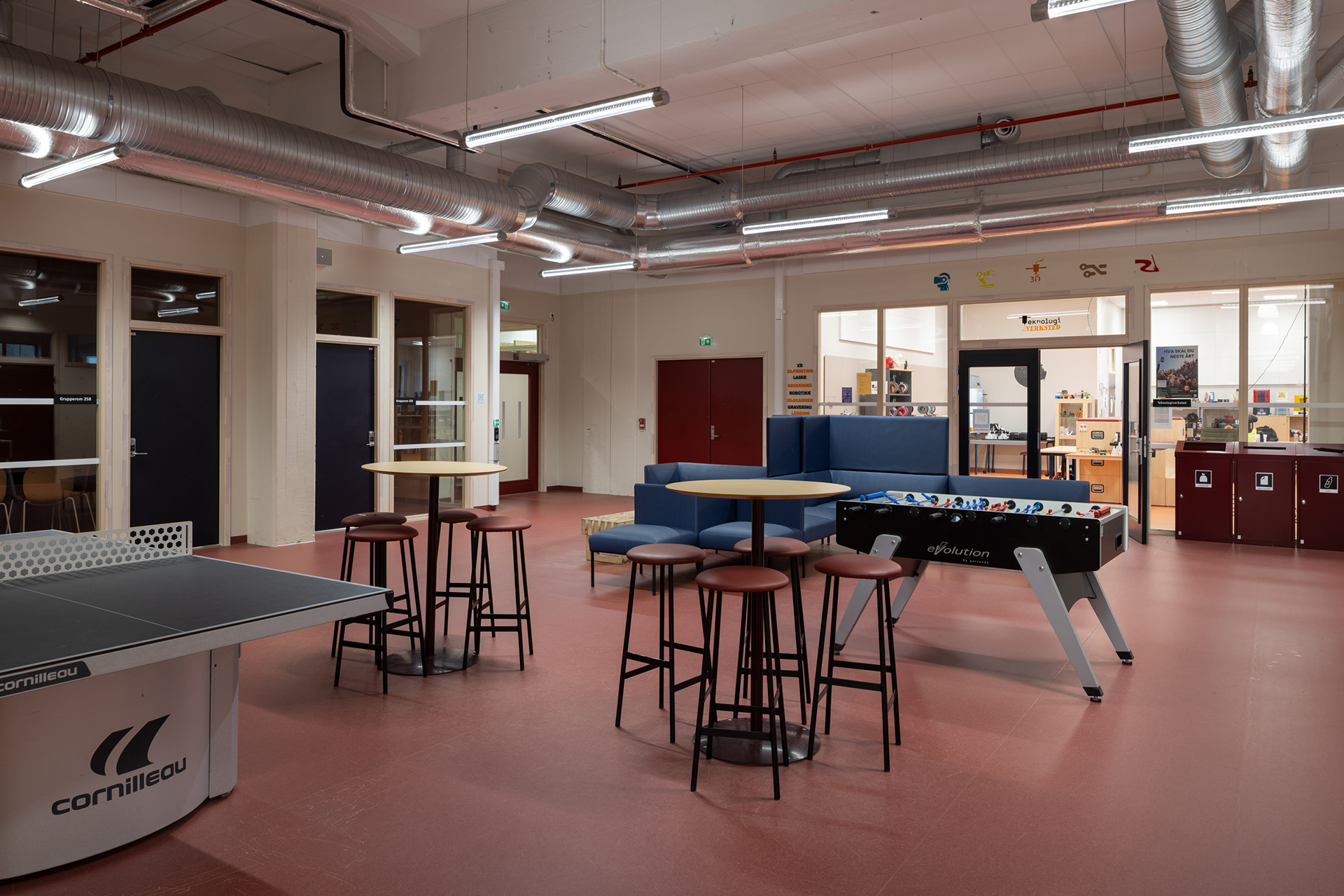 Et oppholdsrom med bordtennisbord, runde bord med krakker, en blå sofa og et bordfotballbord på et rødt gulv.
