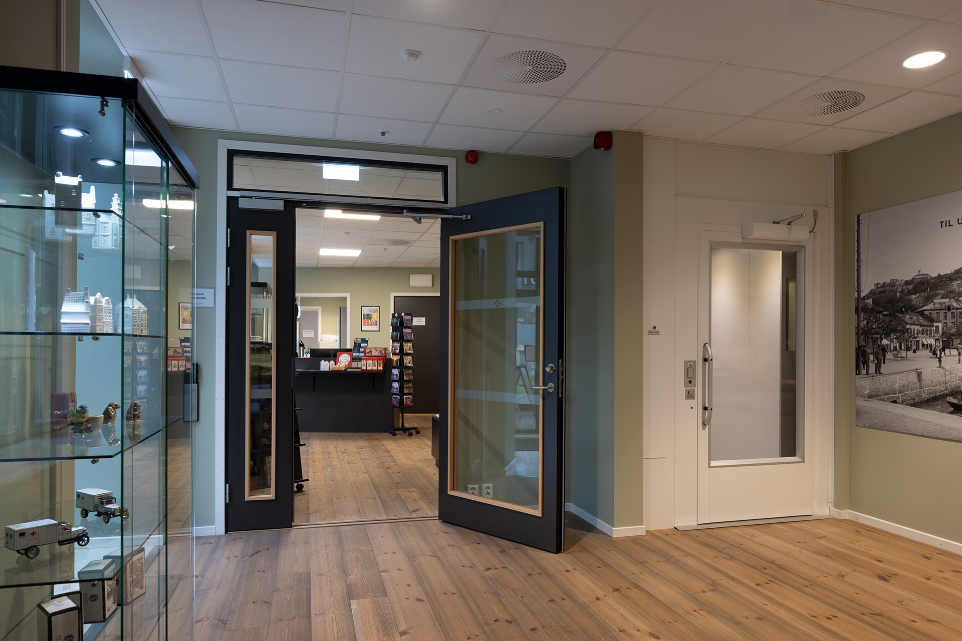 Kameleonen ved parken 8 Et moderne resepsjonsområde med glassmontre til venstre, doble dører som fører til en resepsjon, tregulv og nøytrale veggfarger.