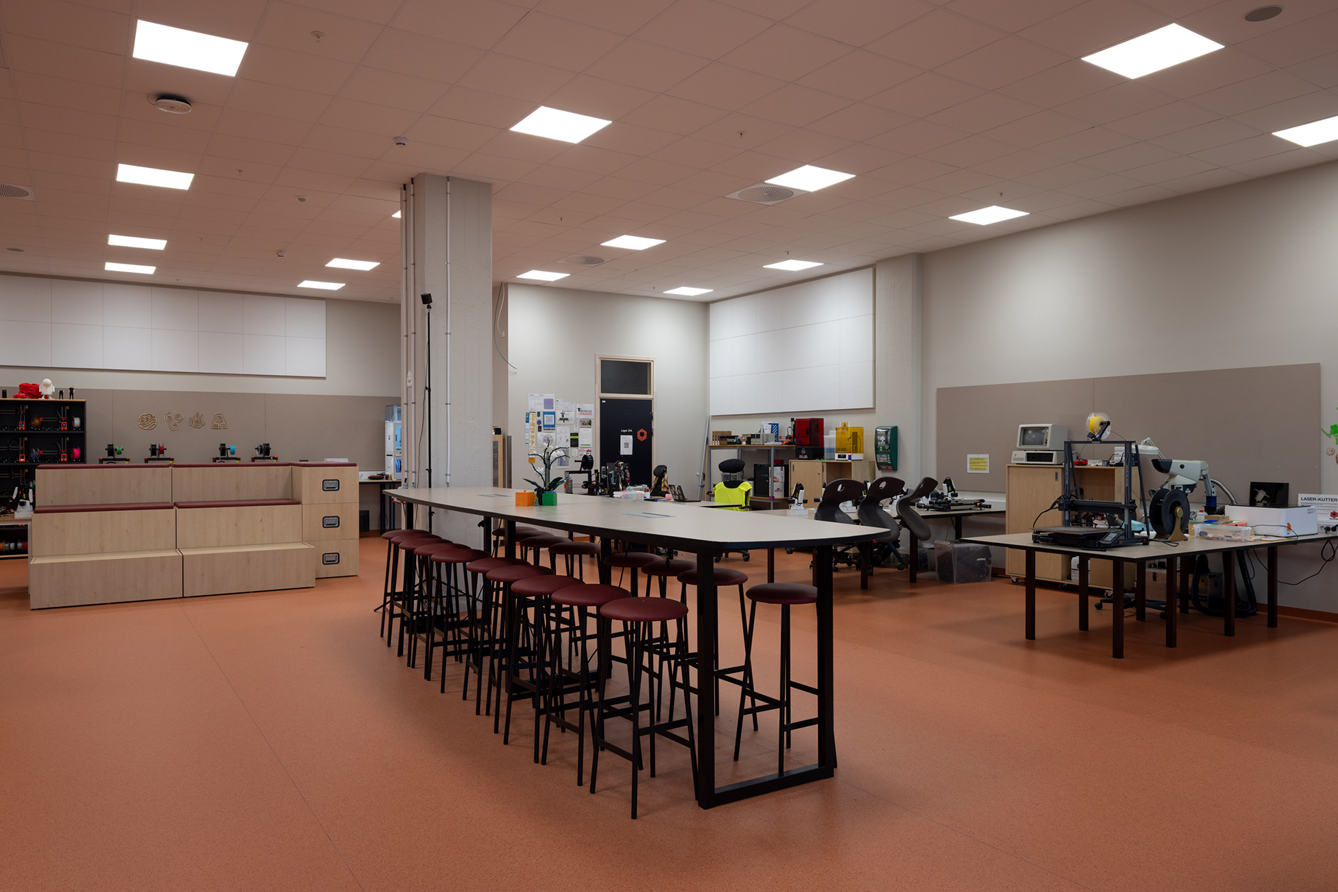 Et moderne verksted med høye bord og krakker, diverse verktøy og utstyr, datamaskiner og hyller, under sterke taklamper.