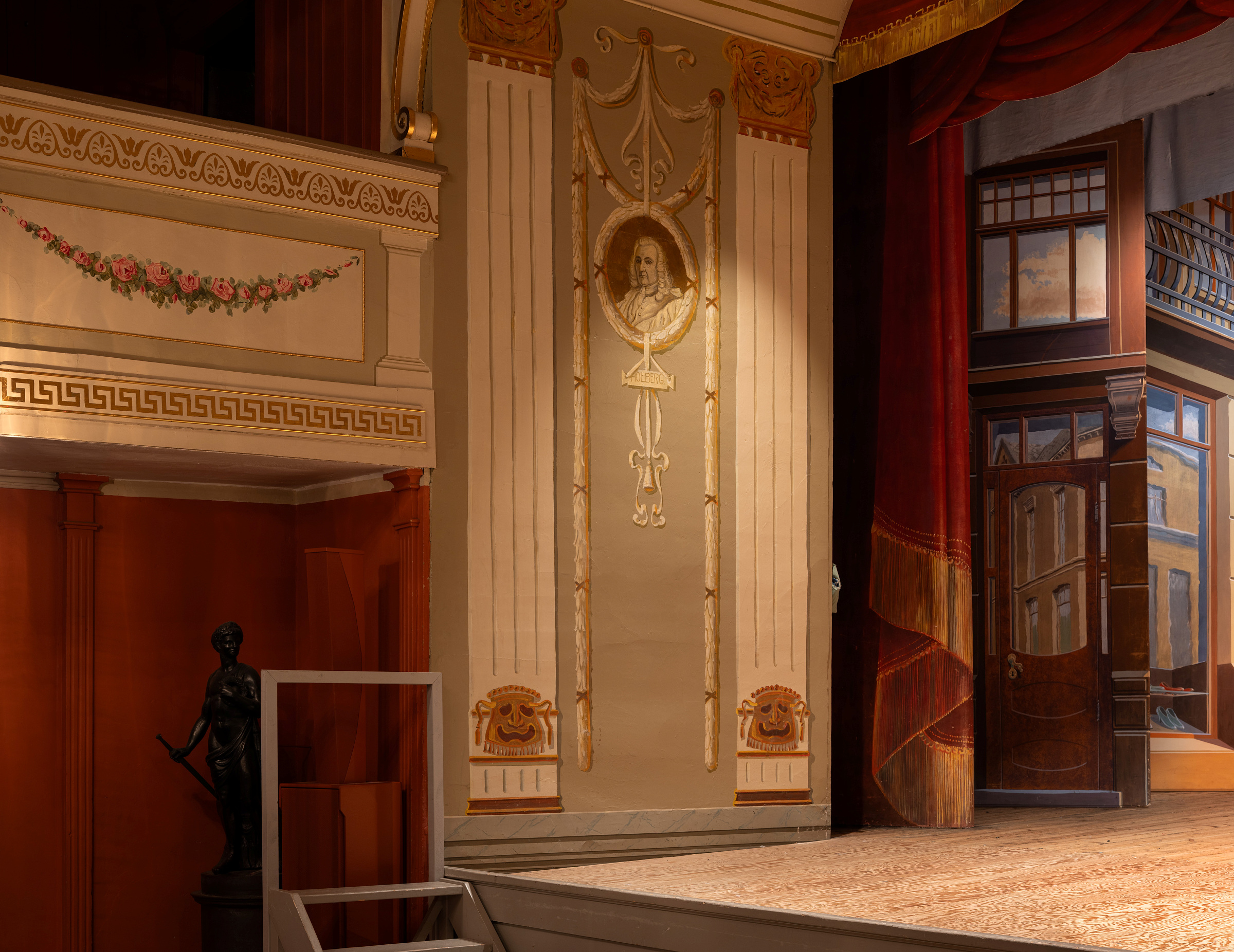 Et utsmykket teaterinteriør med en malt søyle, klassisk bystedekorasjon, statue til venstre og en del av en scene med røde gardiner og et malt bakteppe.