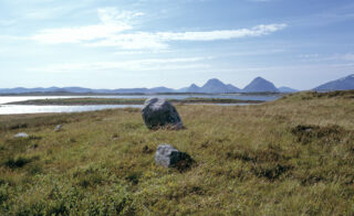 Steinete gressletter nær et rolig vann med fjell i det fjerne under en delvis overskyet himmel.
